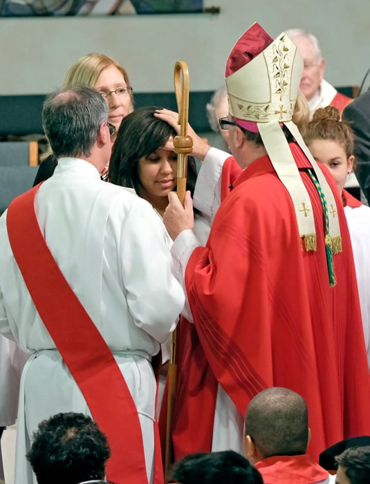 A Bishop confirming a young women with a deacon to his left and a female sponsor behind the confirmandi.